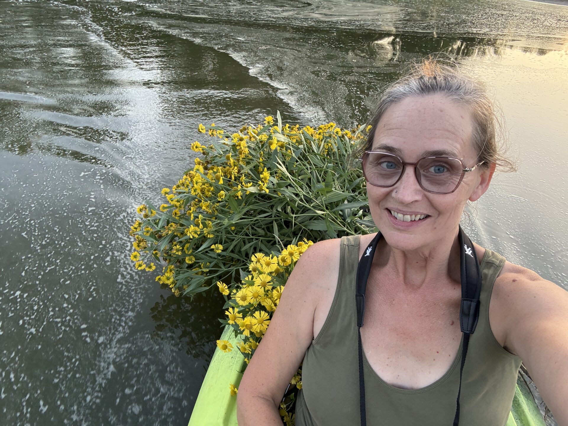 Stephanie smiling in a kayak beside a large bundle of yellow sneezeweed flowers on the Rock River.
