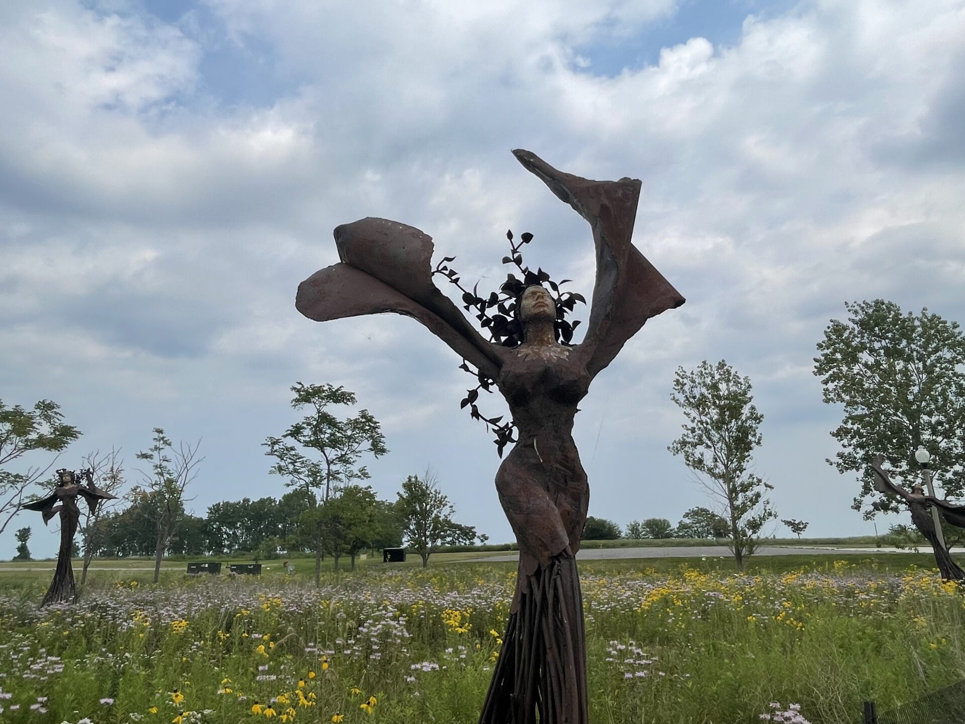 A figure from Dessa Kirk’s Daphne Garden raises leaflike arms above wildflowers at Northerly Island, with two companion sculptures behind it.