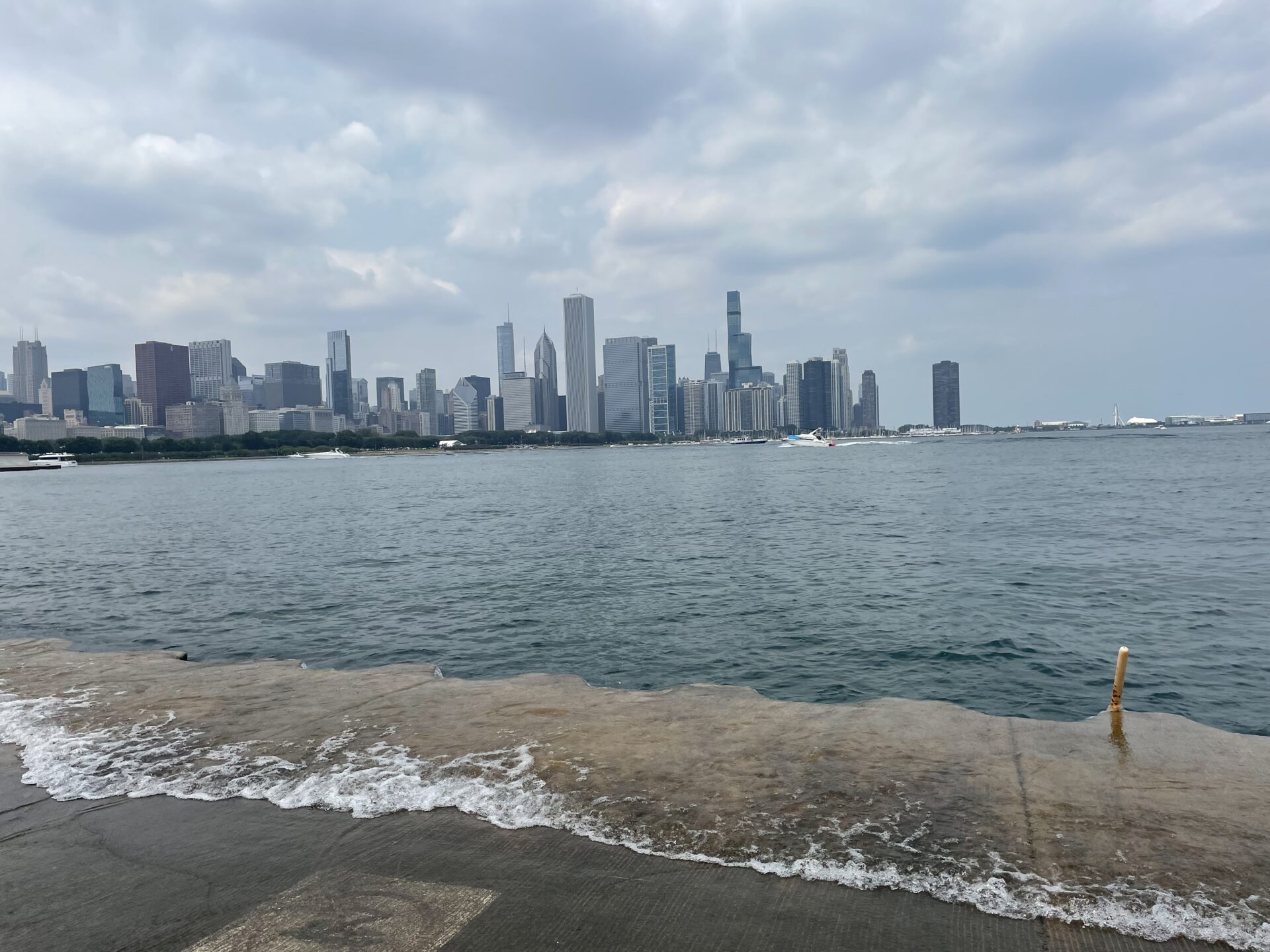 Chicago skyline across Lake Michigan beneath a cloudy sky, with waves washing over the concrete shoreline in the foreground.