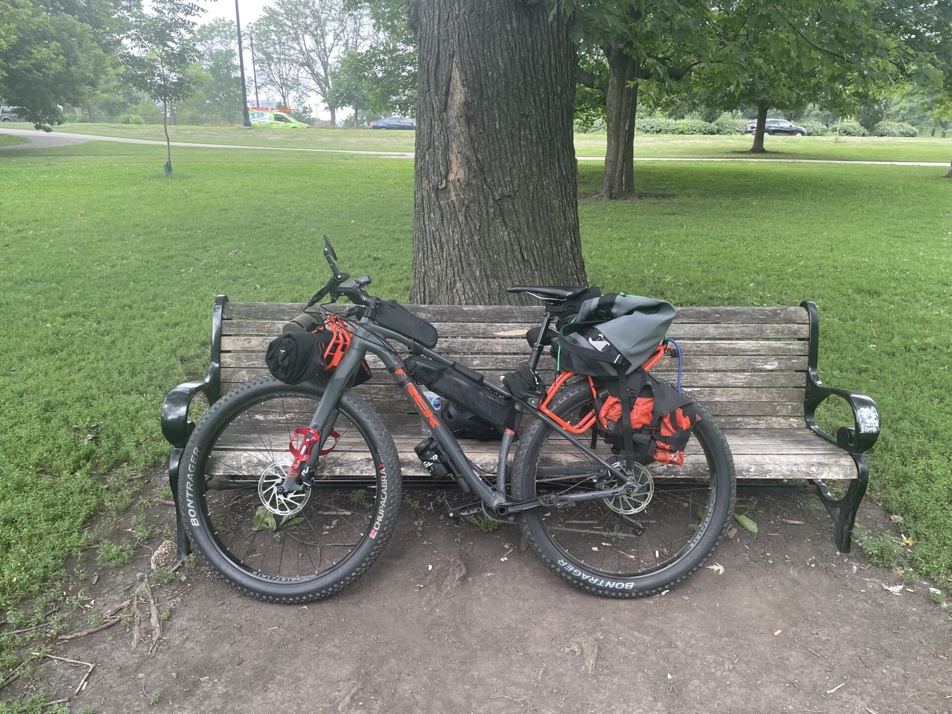 Bicycle loaded with bags leaning against a wooden park bench beneath a large tree.