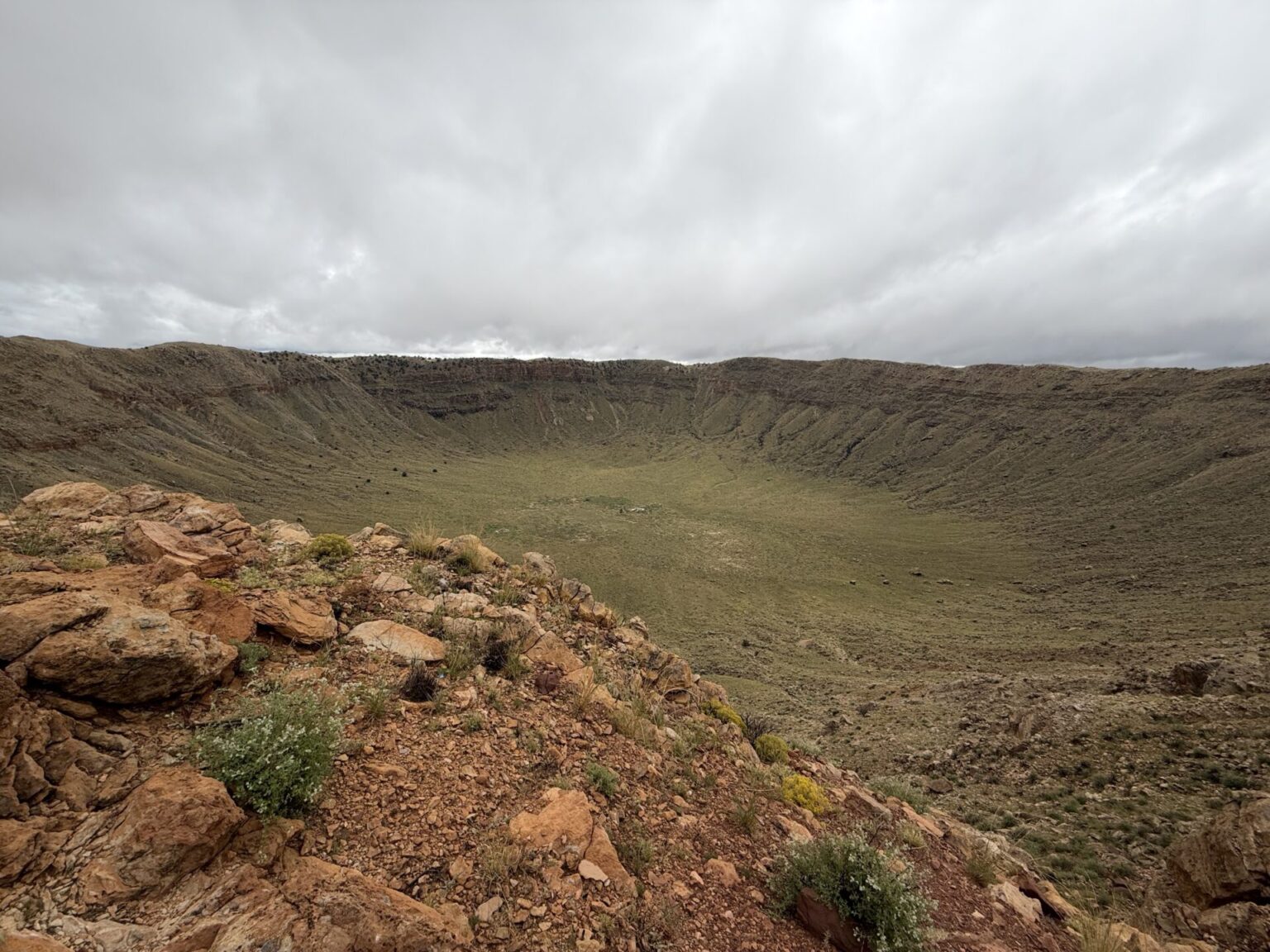 View from the rim of Meteor Crater in Arizona, showing the rocky foreground, crater floor, and surrounding walls beneath a cloudy sky.