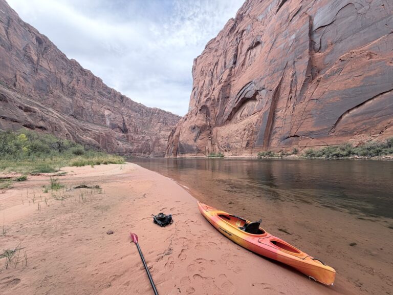 Kayak pulled onto a sandy riverbank beneath towering red sandstone cliffs along the Colorado River in Glen Canyon, Arizona.